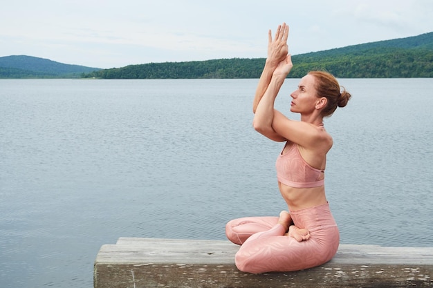 Athletic woman in pink sportswear sitting in lotus position practicing yoga outdoors