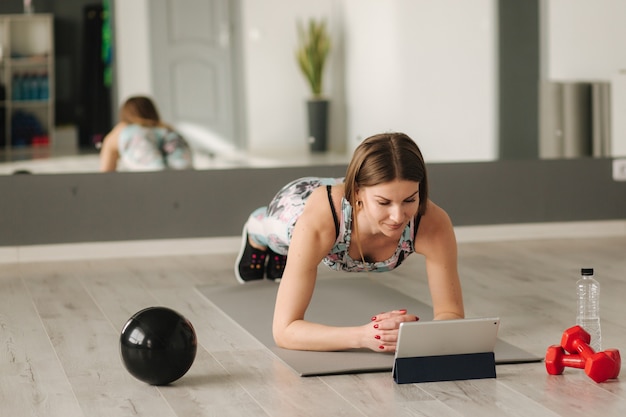 Athletic woman in sportswear does exercises at home. Woman look in tablet while diong exercises