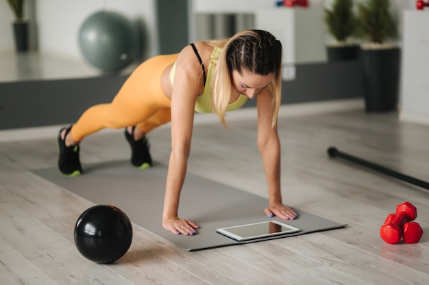 Athletic woman in sportswear does exercises at home. Woman look in tablet while diong exercises.