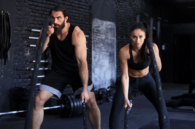 Athletic young couple with battle rope doing exercise in functional training fitness gym.