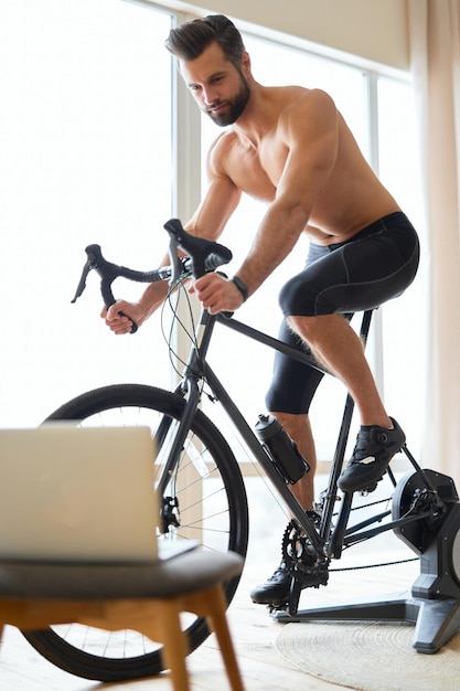 Athletic young man using laptop and riding stationery bike