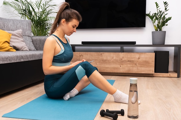 Athletic young woman sitting on a mat relaxing and using a smartphone