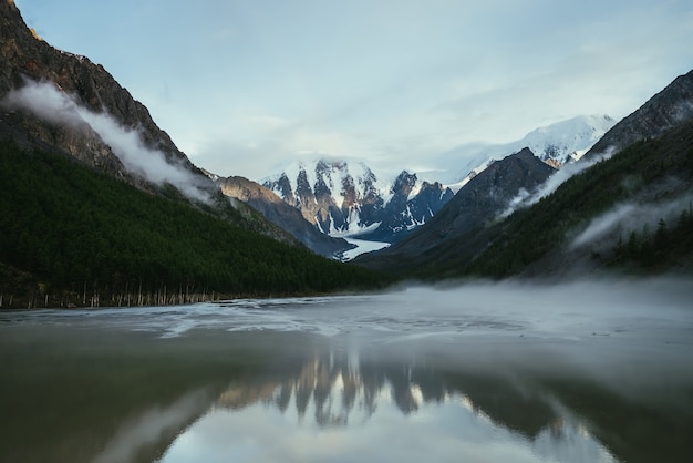 Atmospheric alpine landscape with snowy mountains in golden sunlight reflected on mirror mountain lake in fog among low clouds. Scenic highland scenery with low clouds on rocks and green mirror lake.