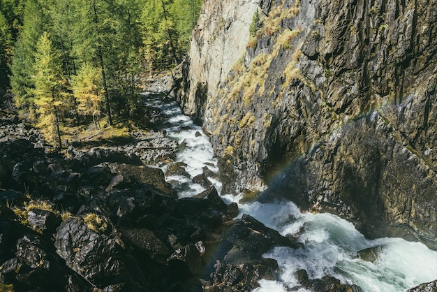 Atmospheric autumn landscape with rainbow above turbulent mountain river among rocks near rocky wall in sunshine. Beautiful alpine scenery with powerful mountain river and autumn forest in sunlight.