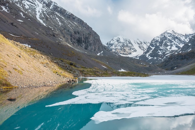 Atmospheric landscape with frozen alpine lake and high snow mountains Sunlit ice floats on transparent water surface of mountain lake Awesome scenery with large snowy mountains reflection in lake