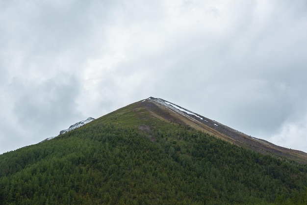 Atmospheric landscape with high green mountain in pyramid shape under gray cloudy sky Somber view to large green forest mountain with bald top with snow in center under rainy clouds at gloomy weather