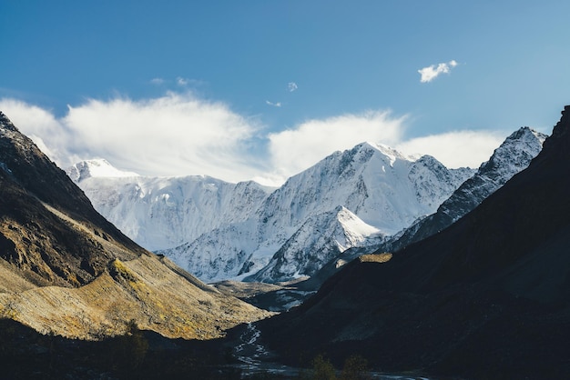 Atmospheric landscape with high snowy mountain with peaked top under cirrus clouds in sky. Low cloud on big snow covered mountain in sunlight. Black rocks in golden sunshine and white-snow pointy peak