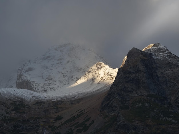 Atmospheric landscape with sharp rocks and high snowy mountain top in blizzard low clouds at overcast Dramatic gloomy scenery with large snow mountains and glacier in gray cloudy sky at sunrise