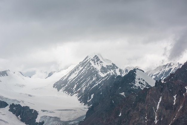 Atmospheric landscape with sharp rocks and high snowy mountain top in rainy low clouds at overcast Dramatic gloomy scenery with large snow mountains and glacier in gray cloudy sky at rainy weather