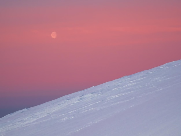 Atmospheric minimalist landscape with moon over high snowy mountain slope in dawn Pink polar dawn sky with moon