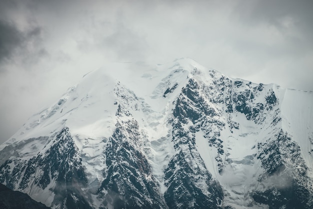 Atmospheric mountains landscape with big snowy mountain top with snow cornices in low clouds. Awesome minimal scenery with snow-white high pinnacle in overcast weather. Giant mountain wall in clouds.