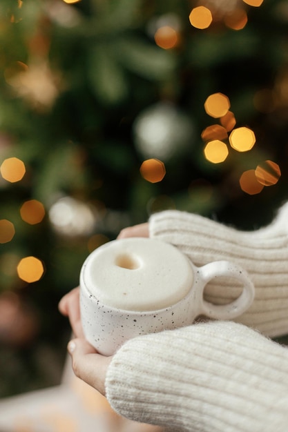 Atmospheric winter hygge Hands holding warm cup of coffee on background of christmas tree with lights Cozy home Woman hands in warm sweater holding stylish mug at illumination bokeh