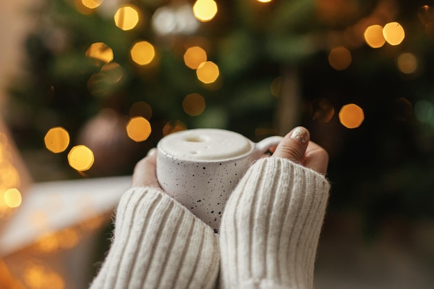 Atmospheric winter hygge Hands holding warm cup of coffee on background of christmas tree with lights Cozy home Woman hands in warm sweater holding stylish mug at illumination bokeh