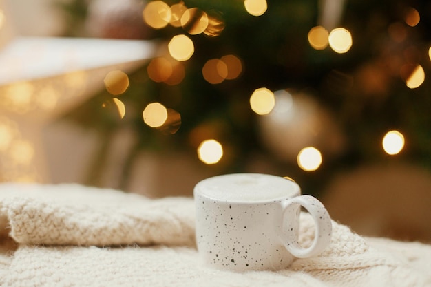 Atmospheric winter hygge Warm cup of coffee on background of christmas tree with lights Cozy home Stylish mug on knitted sweater against golden illumination bokeh Christmas banner