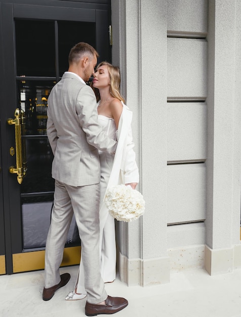 Atractive groom in stylish bright suit hugging young blonde bride woman outdoors Beautiful caucasian girl holding white bouquet of flowers and wears white fashionable wedding suit Wedding style