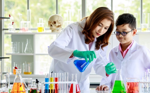 Attentive Asian schoolboy watching female tutor pouring liquid into flasks while conducting experiment with colorful reagents during chemistry lesson in laboratory.