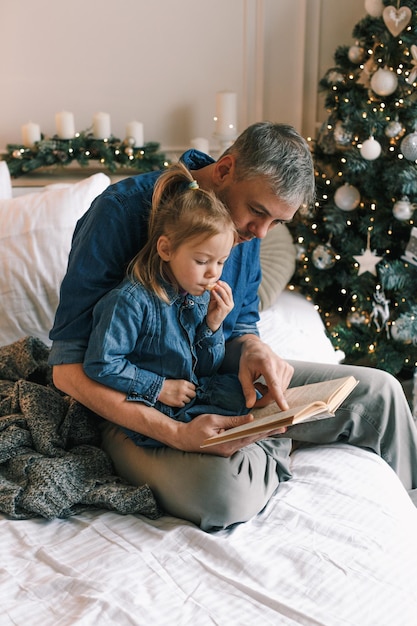 An attentive father reads a Christmas tale for a happy girl sitting on a bed