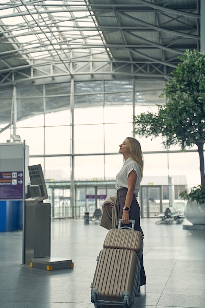 Attentive female person keeping luggage in left hand while being in the airport, standing in semi position