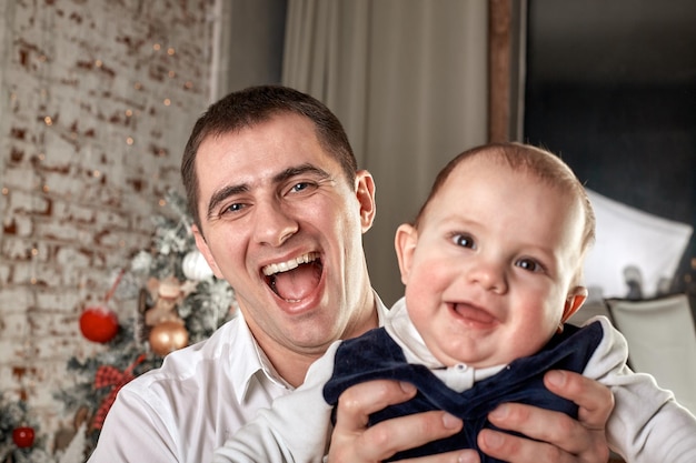 Photo attentive loving father playing with young infant son on white wooden sleigh around a christmas tree with new year gift boxes