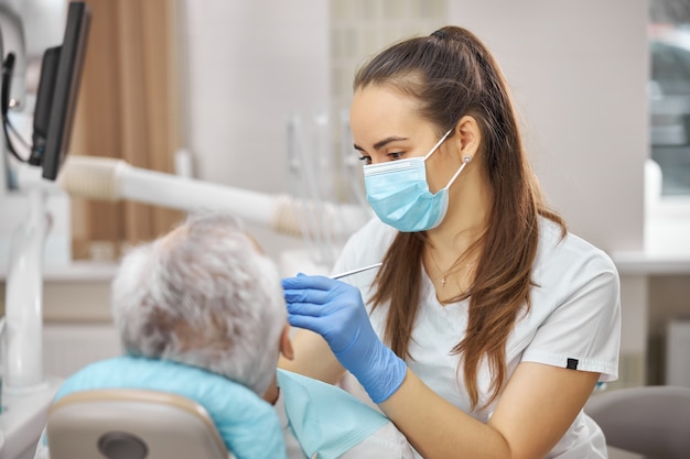 Attentive young female dentist wearing a surgical mask while treating an elderly patient