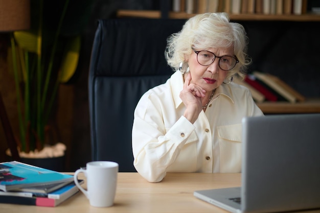 Attentiveness. Mature gray-haired elegant woman in glasses concentrated looking at laptop touching chin sitting at table indoors