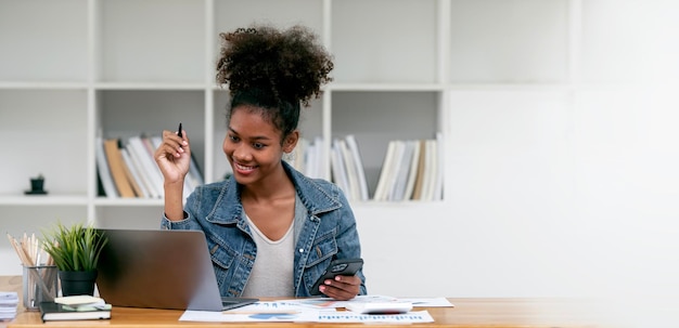 Attractive african young confident businesswoman sitting at the office table working on laptop computer and mobile phone