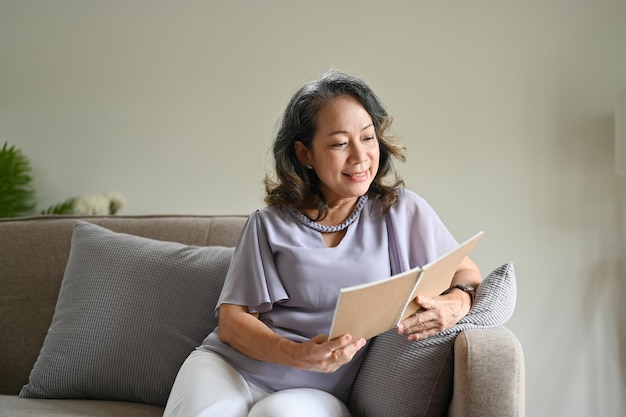 Attractive Asian aged woman relaxing on her sofa in her living room while reading a novel book