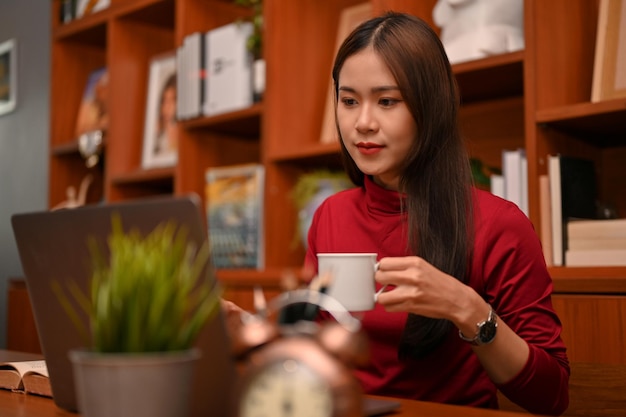 Attractive Asian businesswoman is sipping coffee while working on her laptop computer