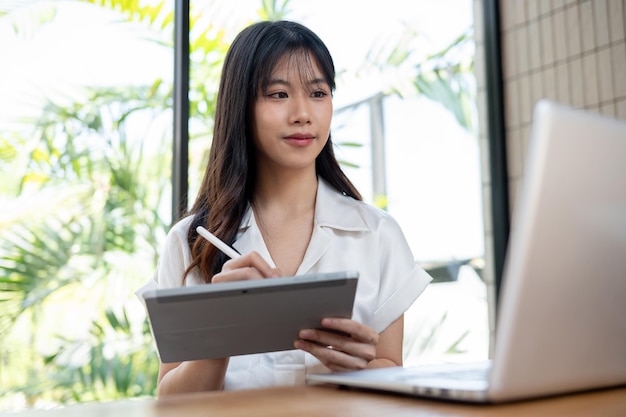 An attractive Asian businesswoman multitasks using her digital tablet and laptop computer in office