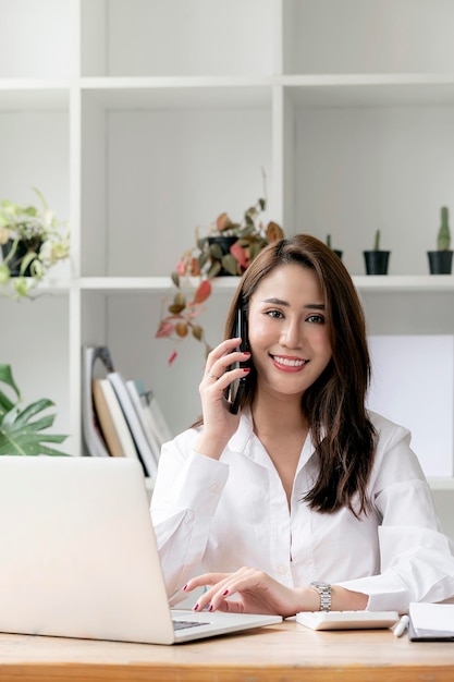 Attractive asian businesswoman using smartphone and working on laptop computer at work, smiling and looking at camera, vertical view.