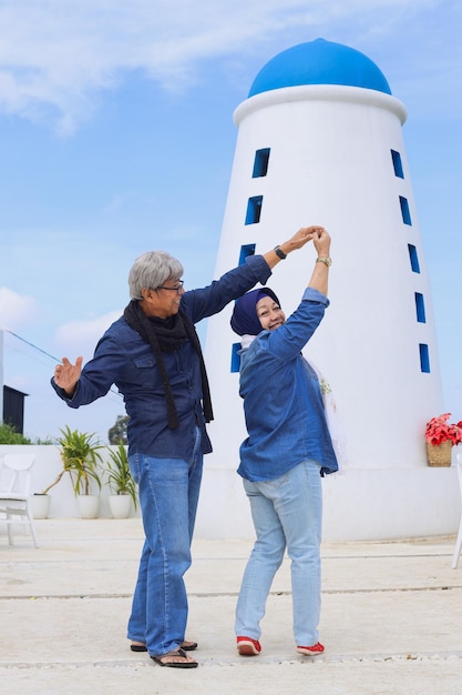 Attractive Asian elderly romantic couple dancing in front of lighthouse with blue sky background