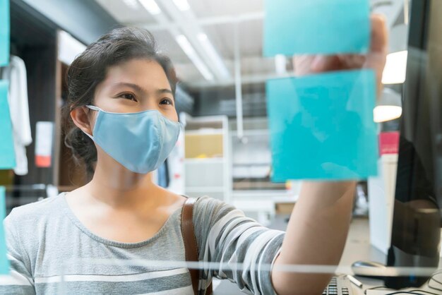 Attractive asian female navy blue sweater working while wearing protective virus mask working in co woking space that have plastic partition social distancing for seat new normal lifestyle