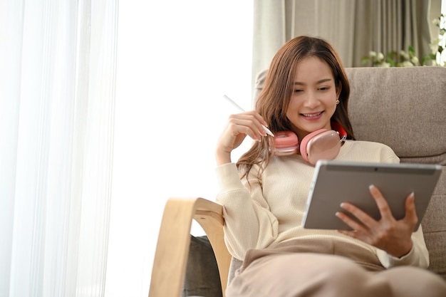 Attractive Asian female with headphones sits on her armchair using a digital tablet touchpad