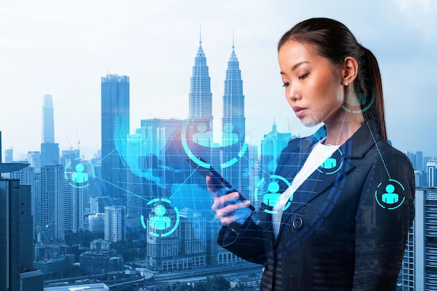 Attractive Asian HR director at international company is looking for highly qualified specialists to recruit by using smartphone Social media hologram icons over Kuala Lumpur