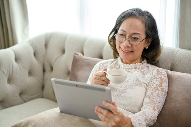 An attractive Asian middleaged woman is sipping coffee while using a tablet in the living room