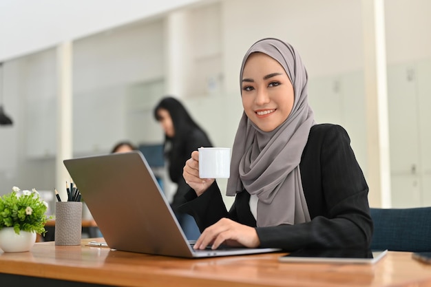 Attractive Asian Muslim businesswoman wearing hijab sipping a morning coffee at office desk