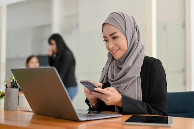 Attractive Asian Muslim businesswoman wearing hijab using smartphone and looking at laptop screen