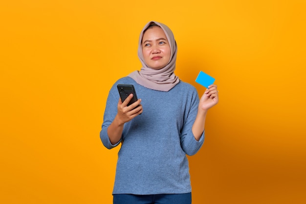 Attractive Asian woman holding mobile phone and showing credit card over yellow background