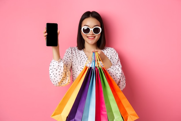 Attractive asian woman showing smartphone app and shopping bags, buying online via application, standing over pink