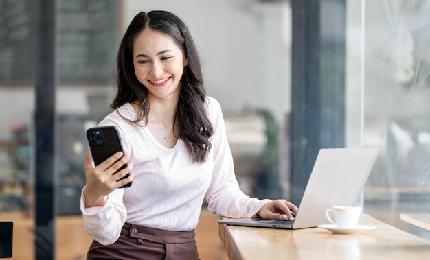 Attractive asian woman smiling and looking at mobile phone in her hand sitting at counter table in cafe with laptop computer