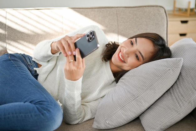 Attractive Asian woman using her smartphone while lying on her comfy couch in her living room