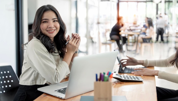 Attractive asian young confident businesswoman sitting at the office table with group of colleagues in the background