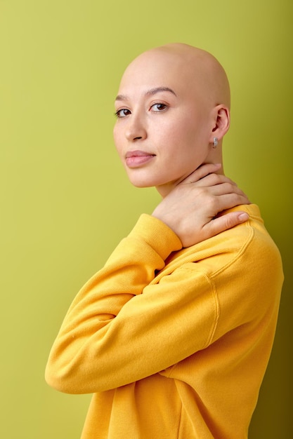 Attractive bald caucasian woman in yellow shirt standing isolated over green studio background, posing at camera, touching neck. beautiful lady with alopecia or cancer. healthcare concept