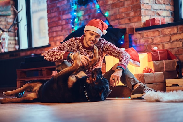 Attractive bearded hipster male sits on a floor with his Rottweiler dog in a room with Christmas decoration.