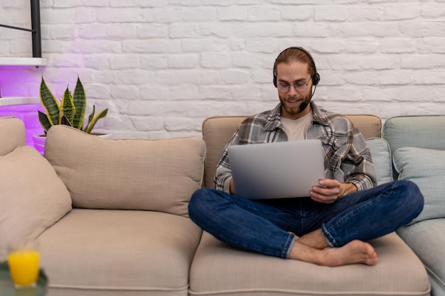 Attractive bearded man gamer playing on laptop at home on cough having online network game using headphones and microphone talking with teammates