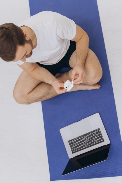 Attractive bearded man holding wireless headphones near laptop and starting morning exercise or yoga