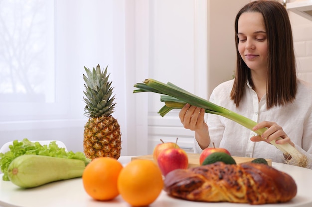 Attractive beautiful Caucasian brown haired woman wearing white shirt sitting at table in kitchen with fruit and vegetables holding leek in hands thinking what to cook