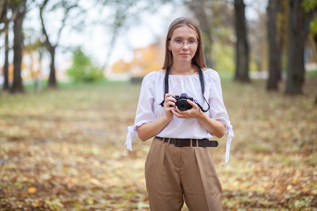 Attractive beautiful young girl holding modern mirror-less camera in autumn park