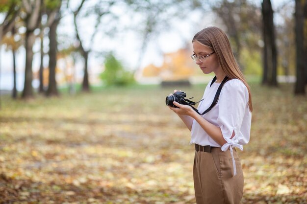 Attractive beautiful young girl holding modern mirror-less camera in autumn park