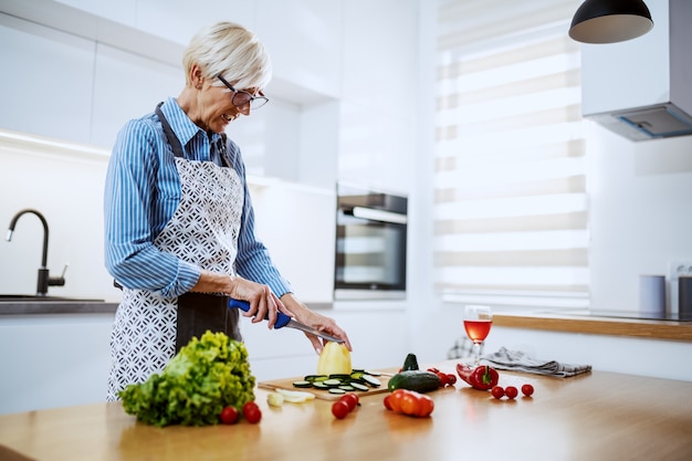 Attractive blond senior woman in apron standing in kitchen at home and cutting yellow pepper